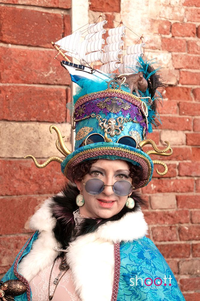 Een persoon draagt ​​een opvallende blauwe steampunk-hoed, versierd met een modelboot, tentakels en paarse accenten, en een blauwe zonnebril.