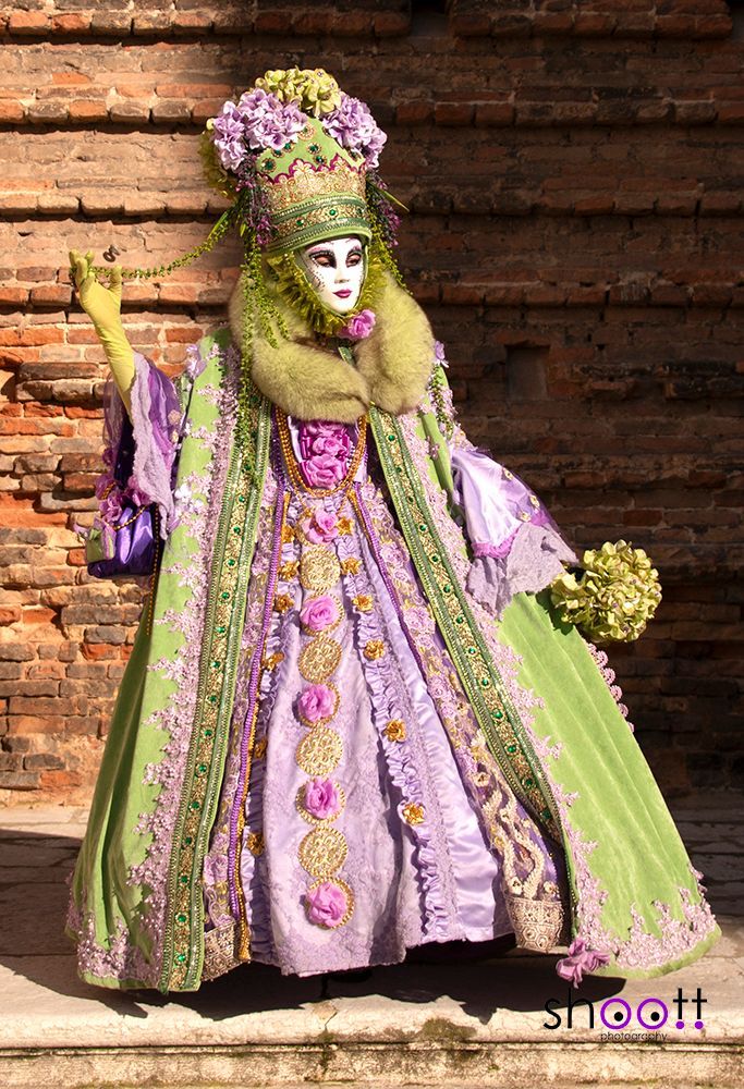 Een persoon in een uitbundig Venetiaans carnavalskostuum met een lila jurk, groene jas, bloemenhoofdtooi en wit masker.