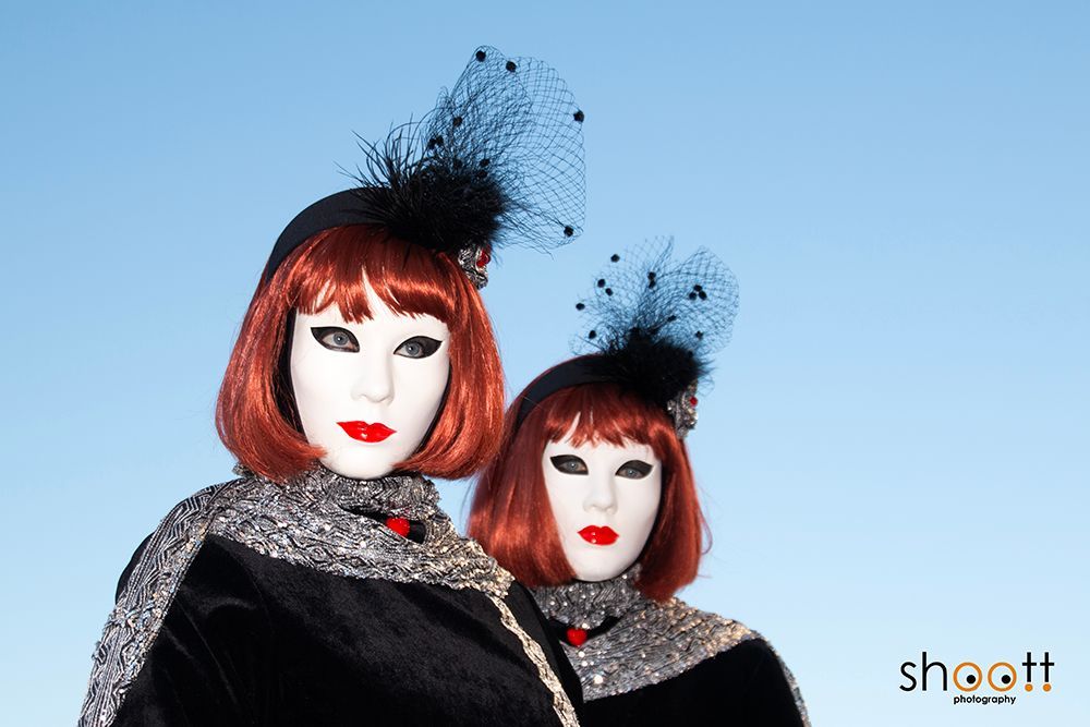 Twee personen met witte maskers, rode bobkapsels en zwarte hoedjes met sluiers, gekleed in zwart-zilveren patronen.