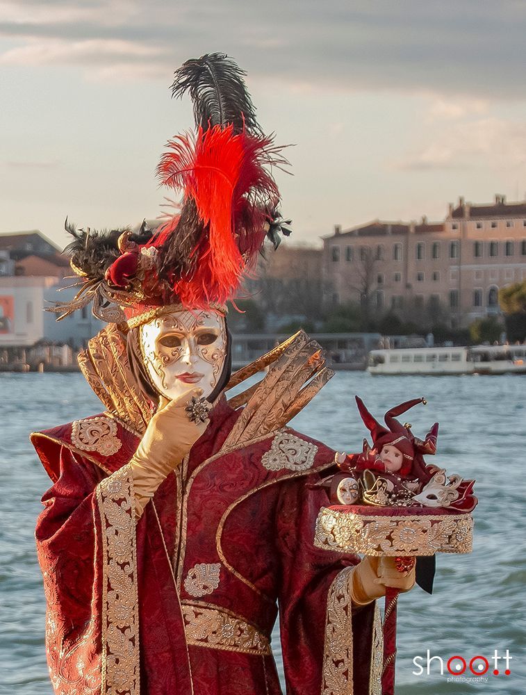 Een artiest die optreedt tijdens het Venetiaanse carnaval, gekleed in een rood, rijk versierd kostuum en masker, houdt een miniatuurpoppetje vast op een dienblad aan het water.