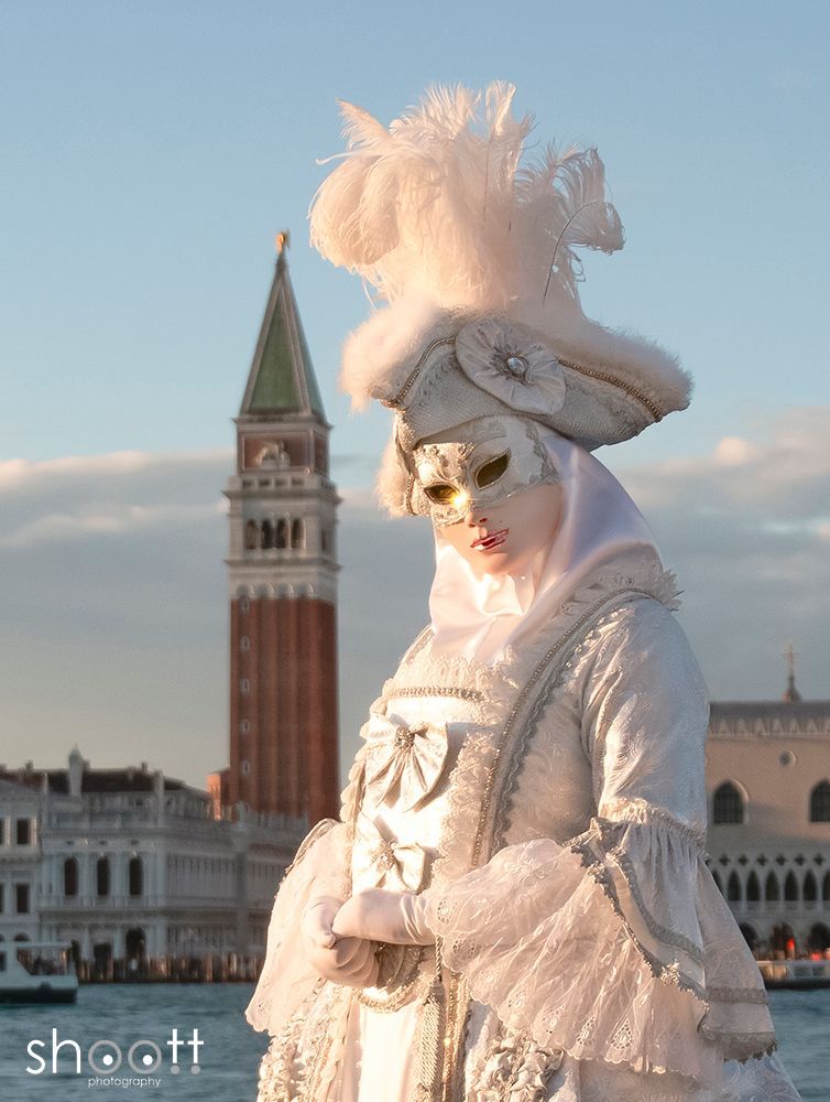 Een persoon in een uitbundig wit Venetiaans carnavalskostuum en masker staat voor de klokkentoren van het San Marcoplein in Venetië.