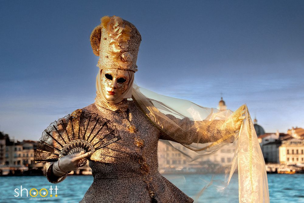 Een persoon met een sierlijk gouden Venetiaans carnavalsmasker, hoed en kostuum, die een decoratieve waaier vasthoudt tegen de stadsachtergrond van Venetië vanaf San Giorgio.