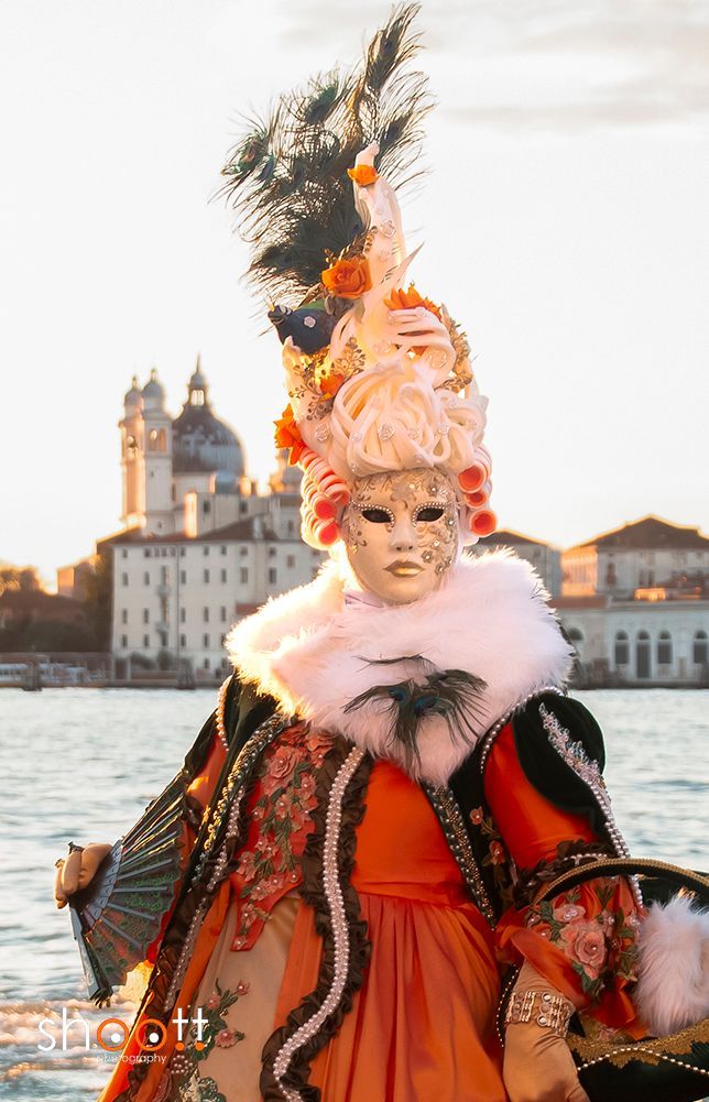 Een Venetiaans carnavalskostuum met een grote pruik van veren, een sierlijk masker en een oranje jurk, staand aan de waterkant van het San Marcoplein in Venetië.