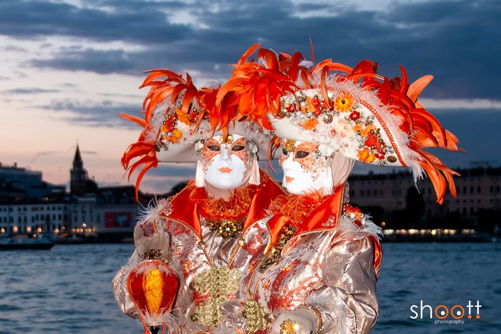 Twee personen, gehuld in sierlijke oranje en gouden Venetiaanse maskers en kostuums, staan ​​voor het stadsgezicht van Venetië aan het water bij schemering.