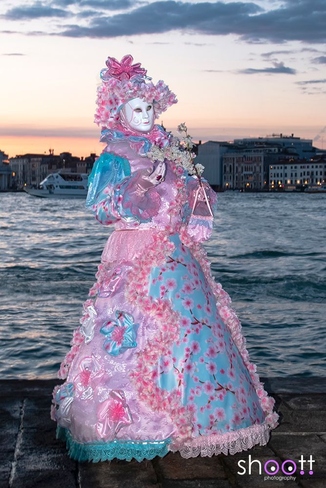 Een gemaskerde figuur in een uitbundige roze en blauwe jurk met ruches staat bij zonsondergang aan het water in Venetië bij ondergaande zon.