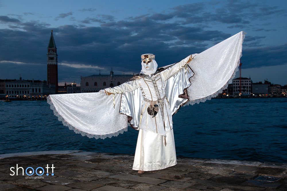 Een gemaskerde figuur in een uitgebreid wit kostuum met grote, vleugelachtige mouwen staat bij het water in Venetië in de schemering.