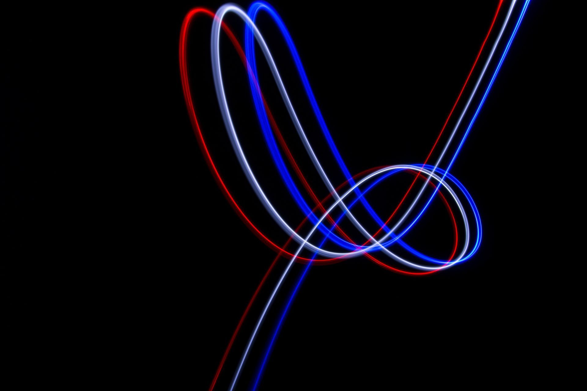 Een lichtschilderij op een zwarte achtergrond met gebogen rode, witte en blauwe lichtsporen die een lusvormige, abstracte figuur vormen, ook wel lightpainting genoemd.