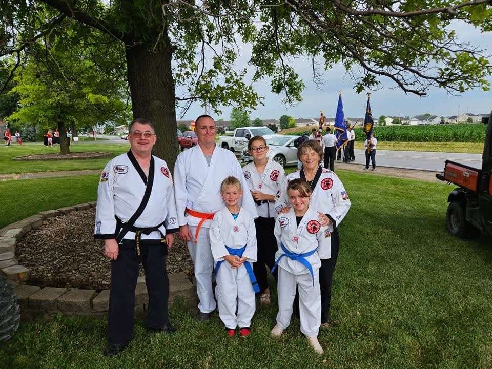 Six people in martial arts uniforms posing outdoors by a tree, with other people and flags in the background.