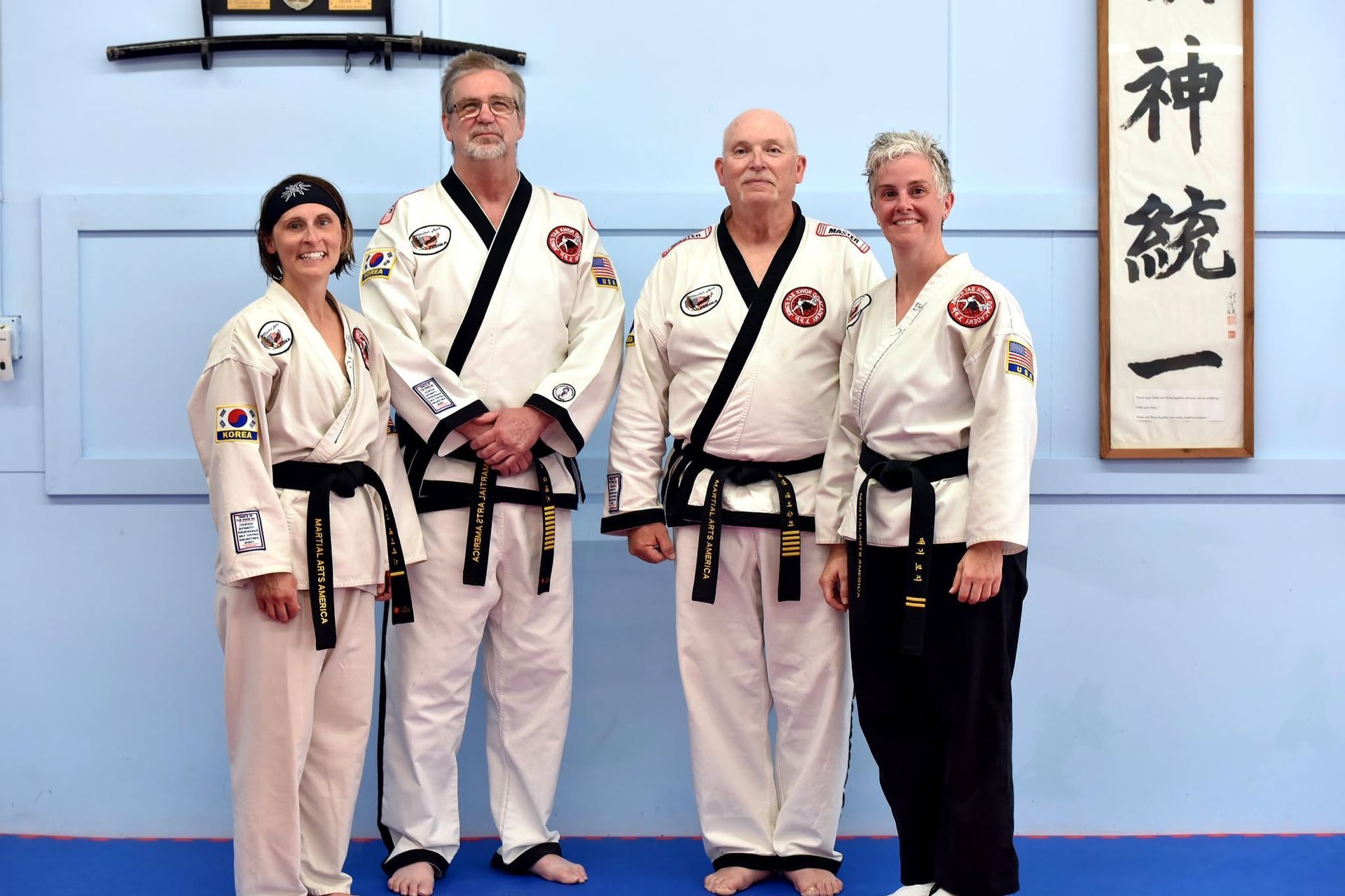 Four martial arts practitioners in white uniforms pose indoors beside a wall scroll.