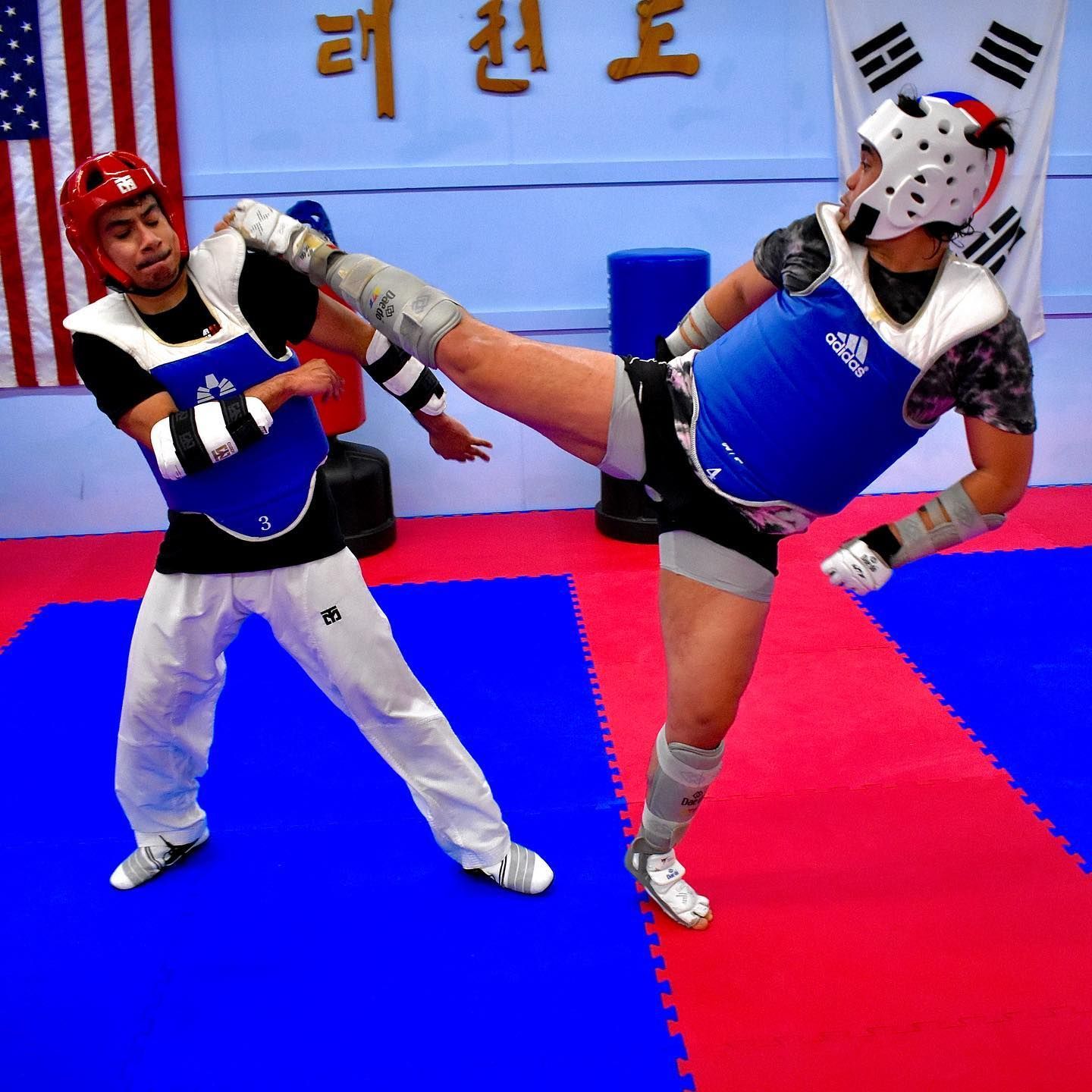 Two martial artists spar on red and blue mats, one delivering a high kick in a training gym.