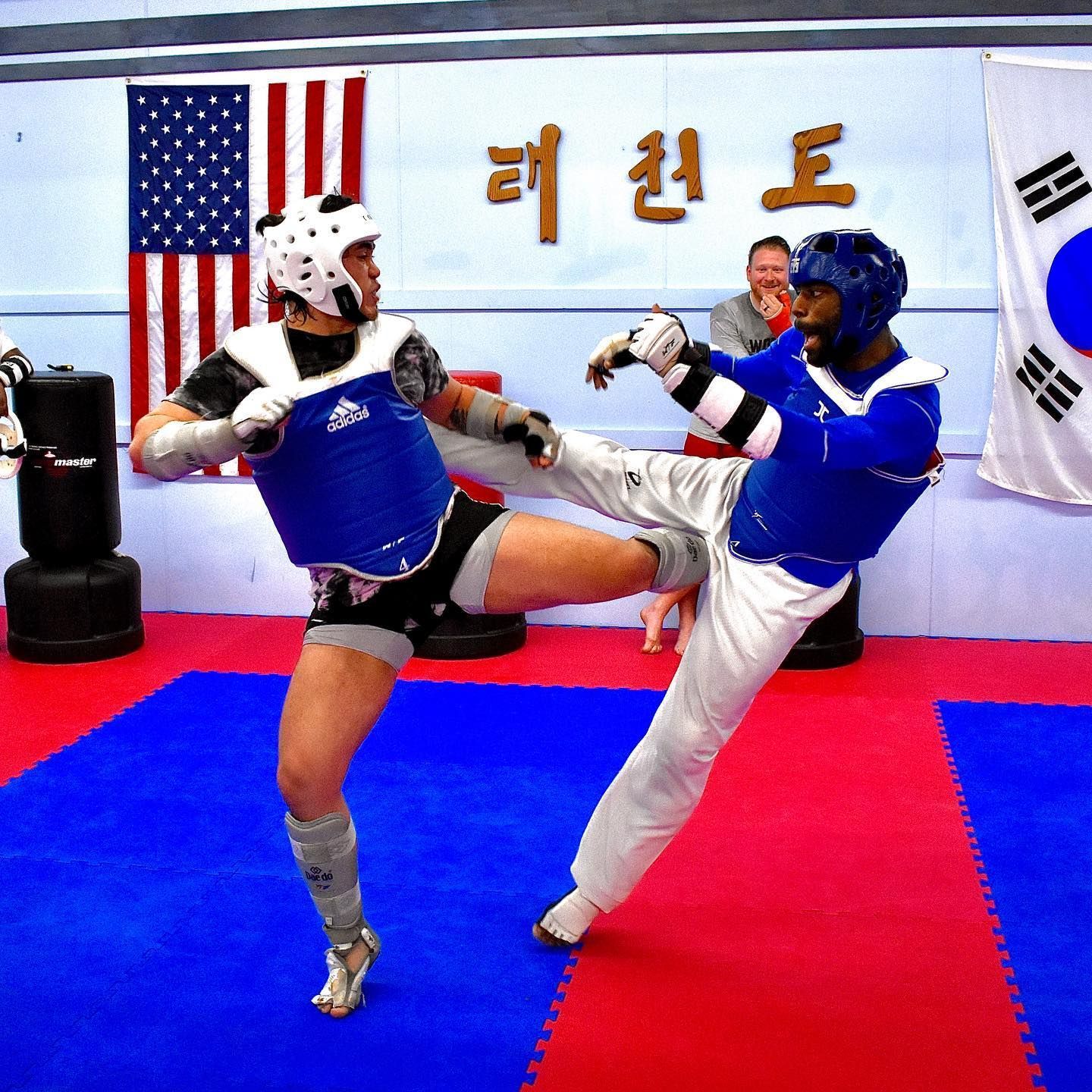 Two martial artists sparring with kicks in a taekwondo studio, with flags on the wall.
