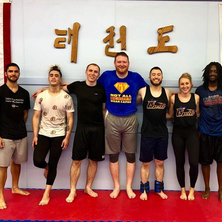 Seven people standing in a martial arts dojo, posing in front of a wall with Korean lettering.