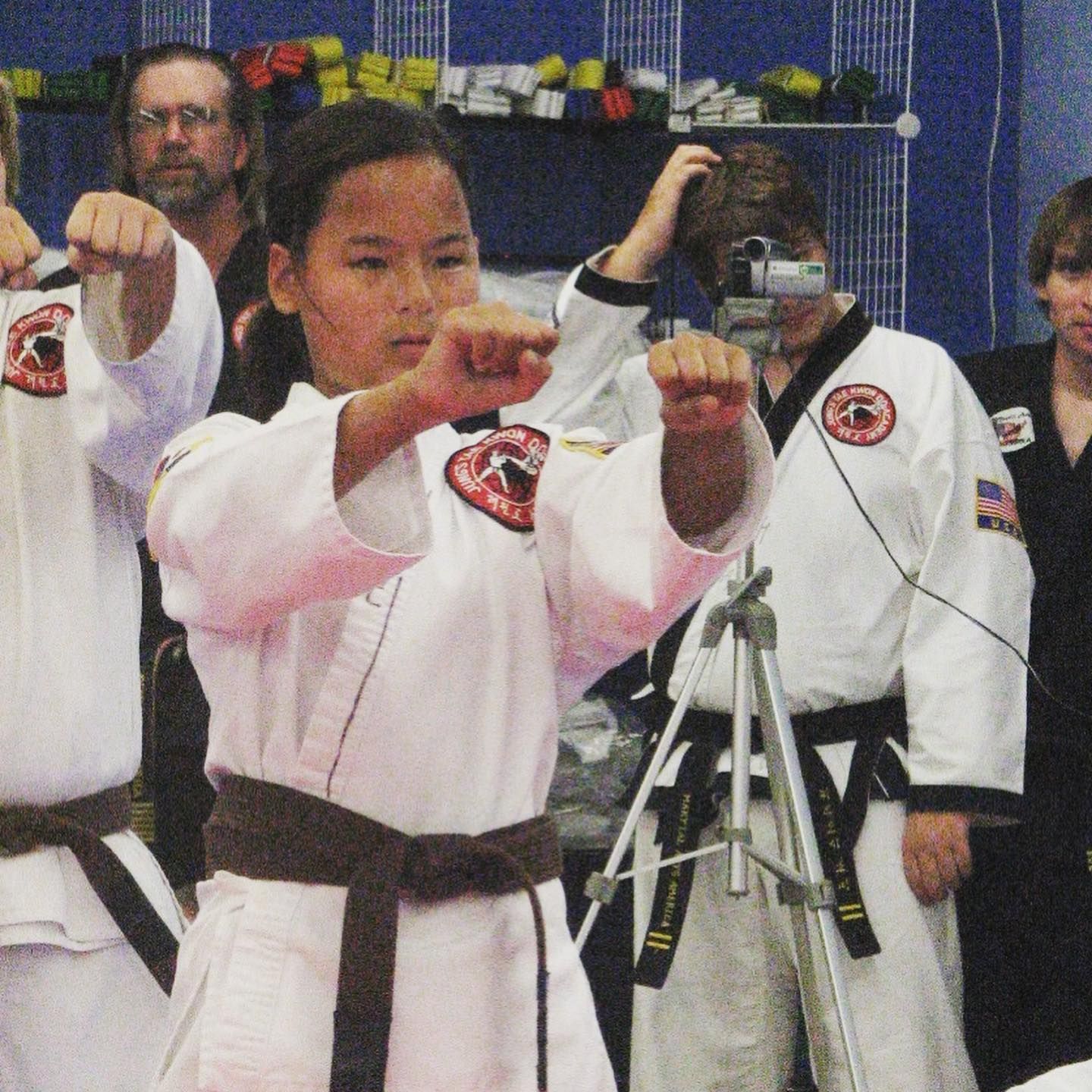 Martial arts students in white uniforms practice punching in a studio, with a tripod and observers behind them
