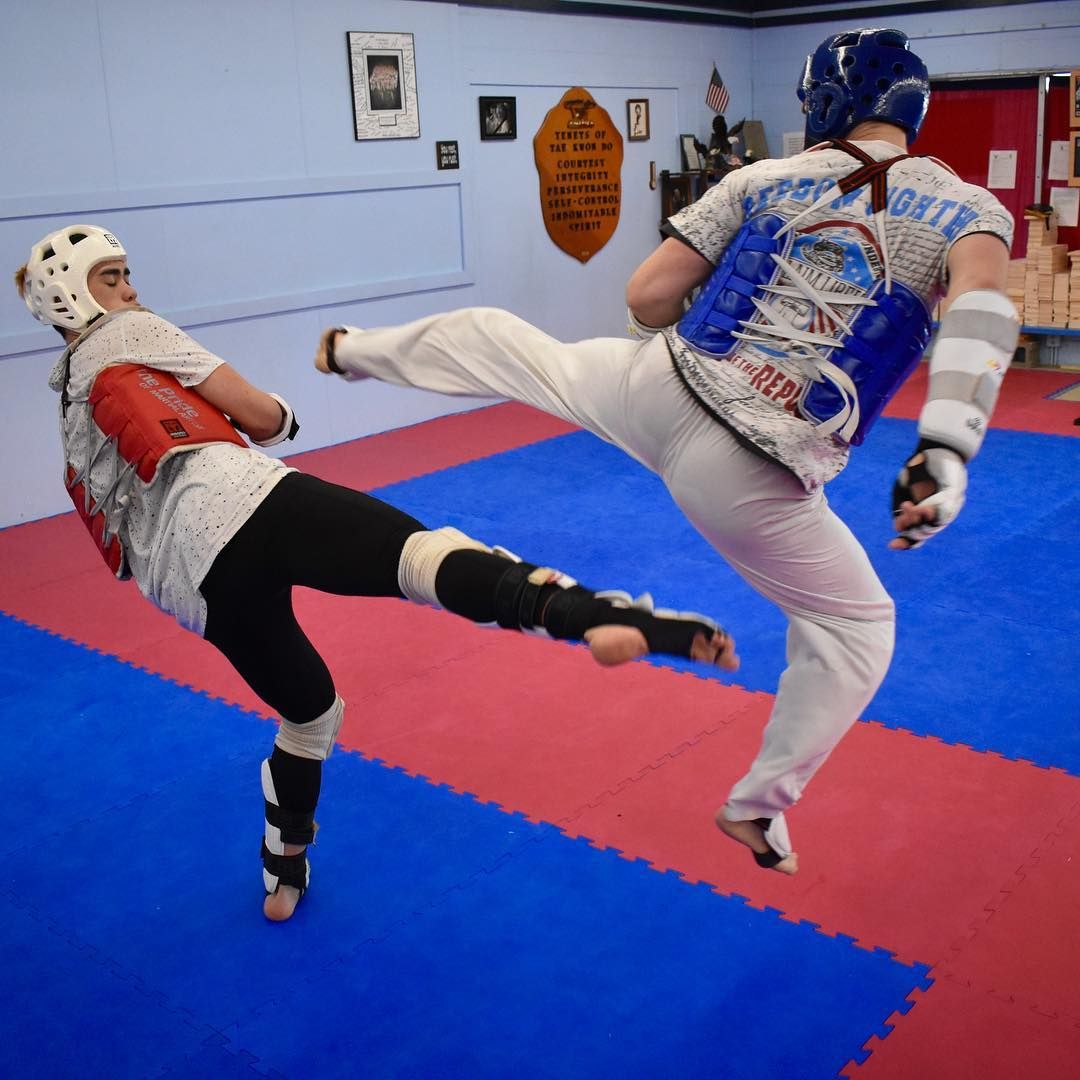 Two martial artists sparring in a gym, both mid-kick on red and blue mats.