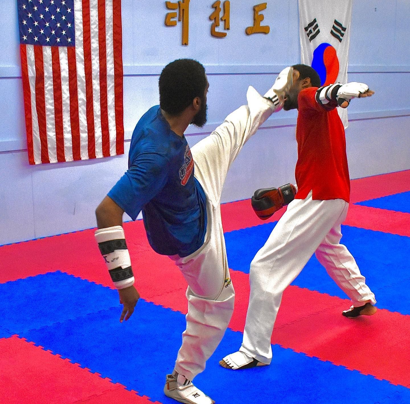 Two martial artists spar on red-and-blue mats in a taekwondo studio with flags on the wall.