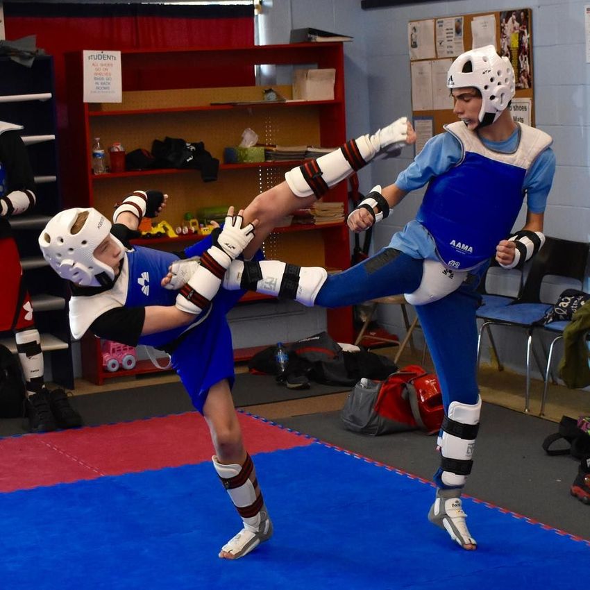 Two martial artists in blue sparring gear practice high kicks on mats in a training room