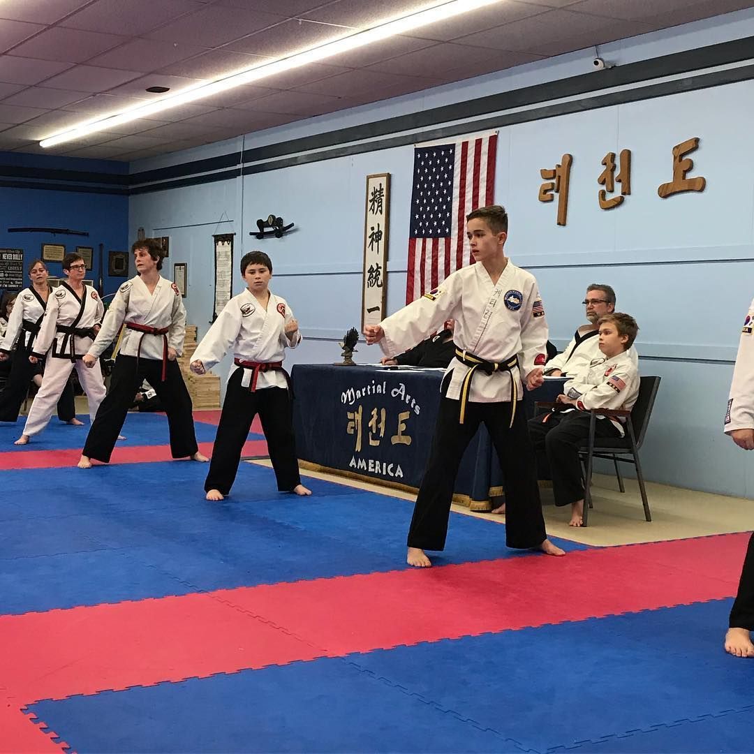 Martial arts class practicing in a dojang, students in white uniforms on blue mats with an American flag and Korean text.