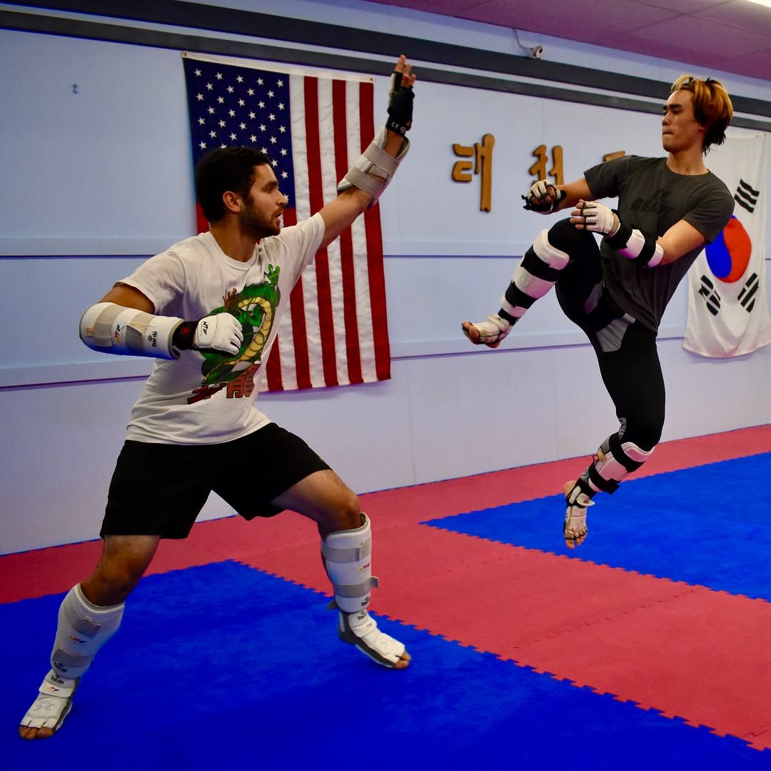 Two martial artists spar in a gym, one lunging with raised guard as the other kicks midair.