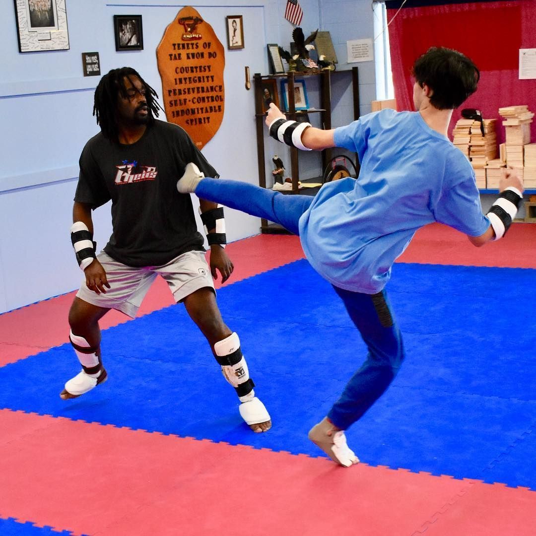 Two martial artists sparring in a dojo, one kicking while the other blocks on blue and red mats.
