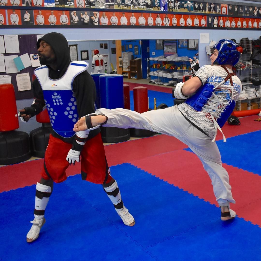 Two martial artists spar in protective gear, one mid-kick in an indoor training gym.