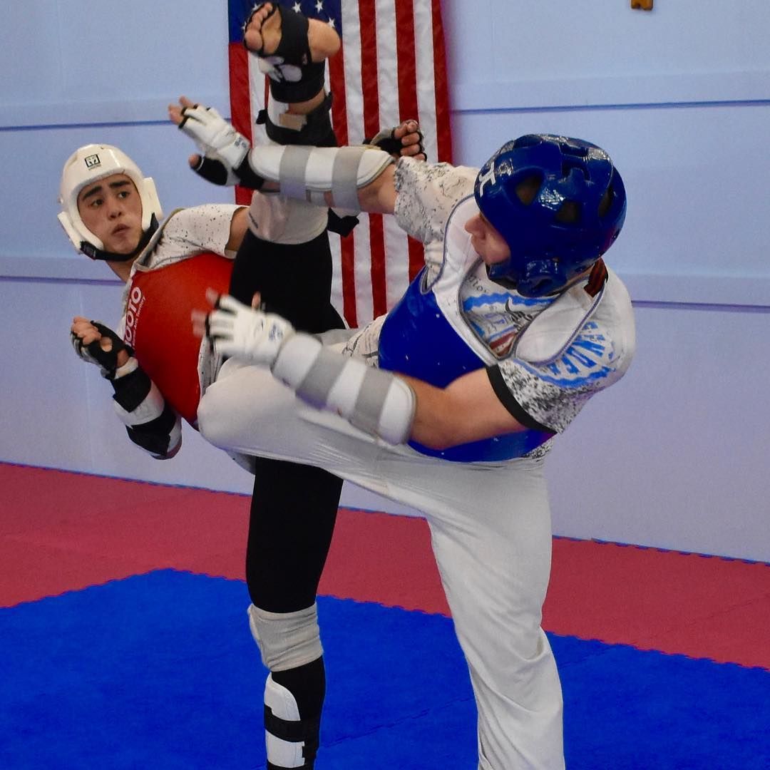 Two martial artists sparring indoors, one in blue landing a high kick on an opponent in red protective gear