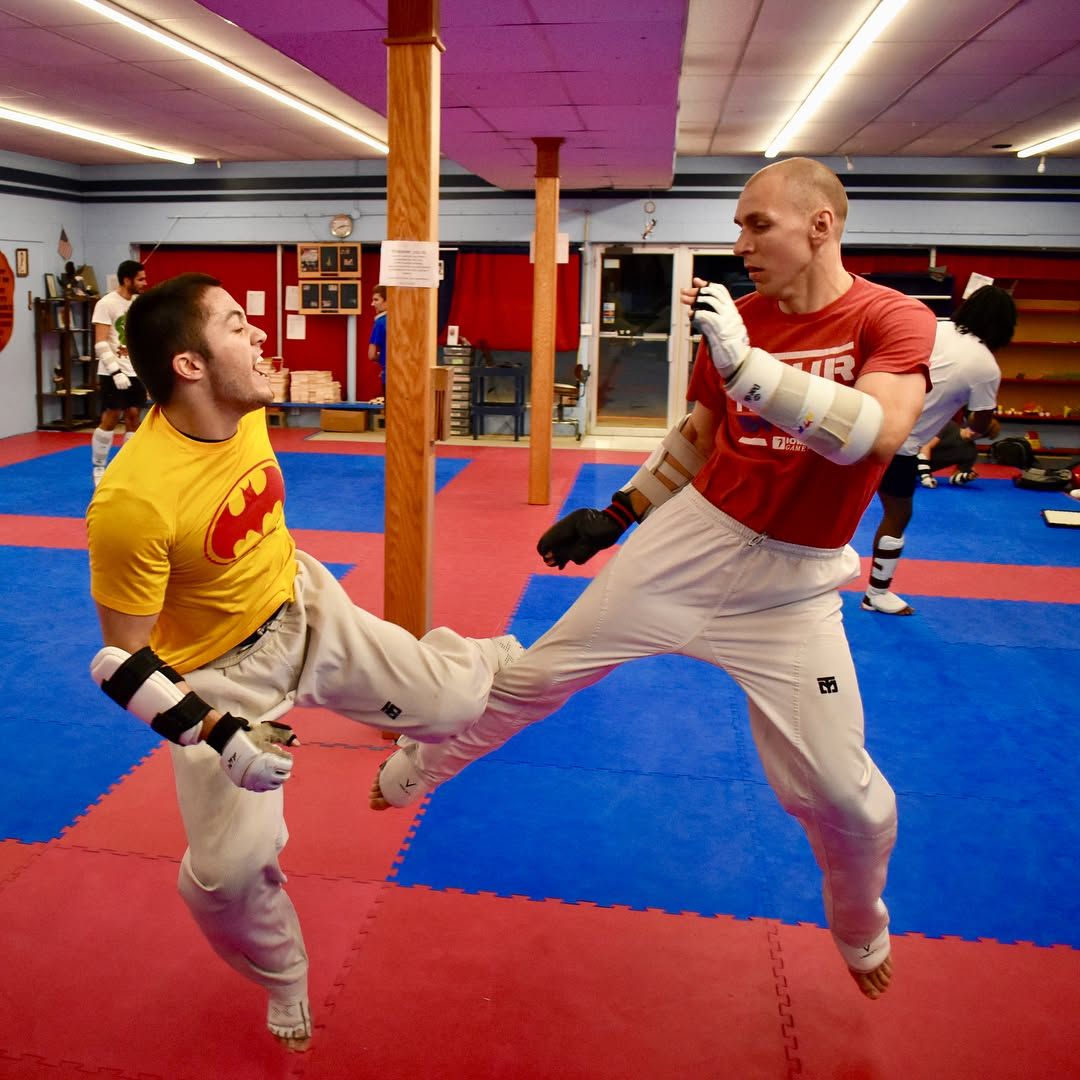 Two martial artists spar in a gym, one in a yellow shirt kicking a red-shirted opponent.