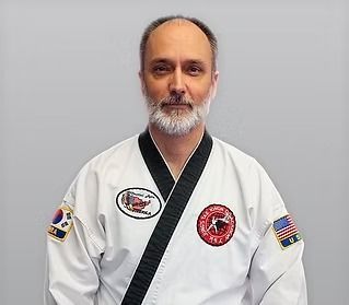 Man in a white martial arts uniform with black trim and patches, standing against a plain gray background