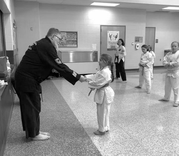 Martial arts instructor handing a belt to a student in a dojo, with other students watching nearby