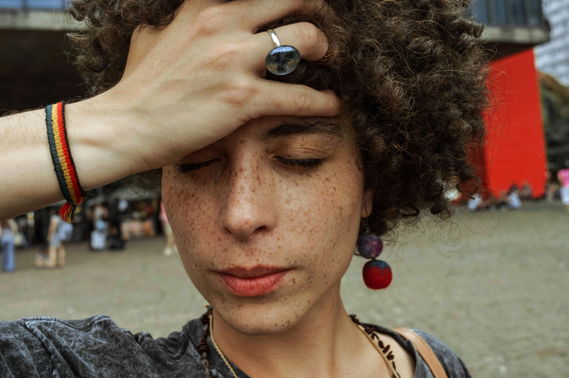 Person with eyes closed and hand on forehead at an outdoor gathering, wearing a blue ring and striped bracelet