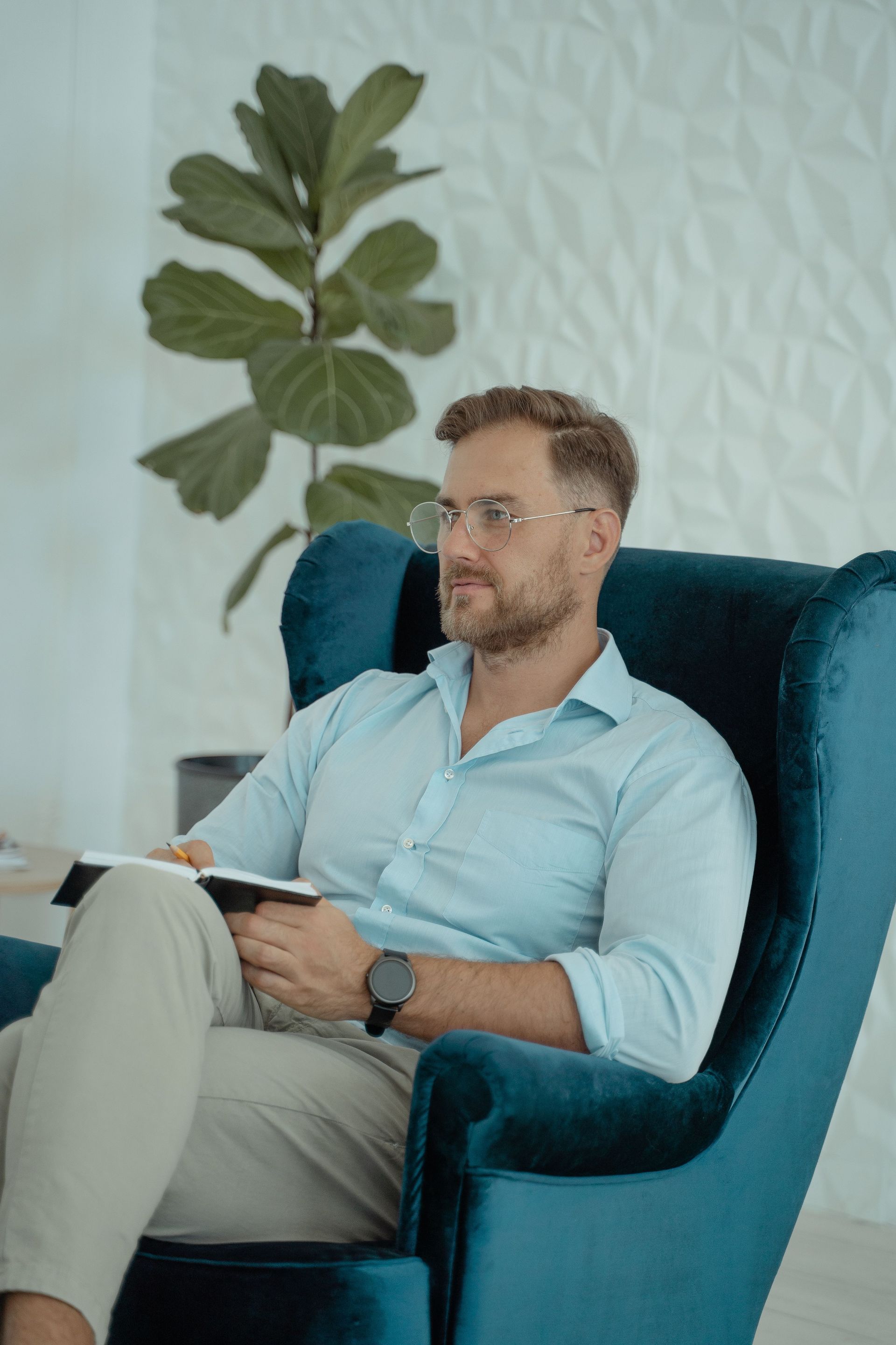 Man sitting in a teal armchair, reading a notebook in a bright room with a large plant behind him.