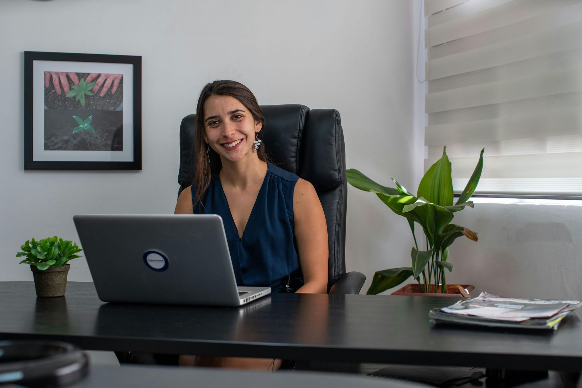 Woman smiling at desk with laptop in a modern office, framed art and potted plants behind her