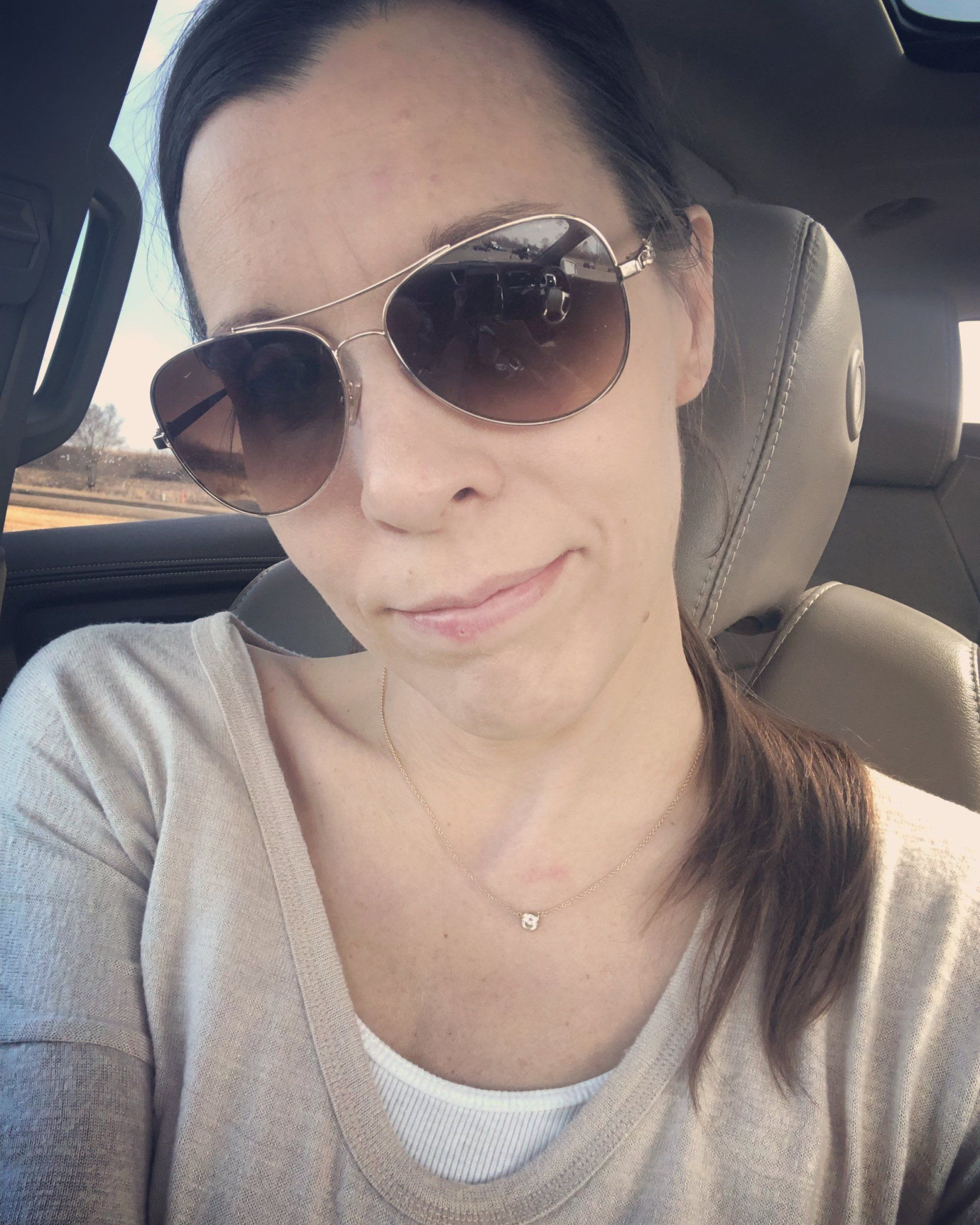 amy in car with hair in pony tail and sunglasses on. No expression.