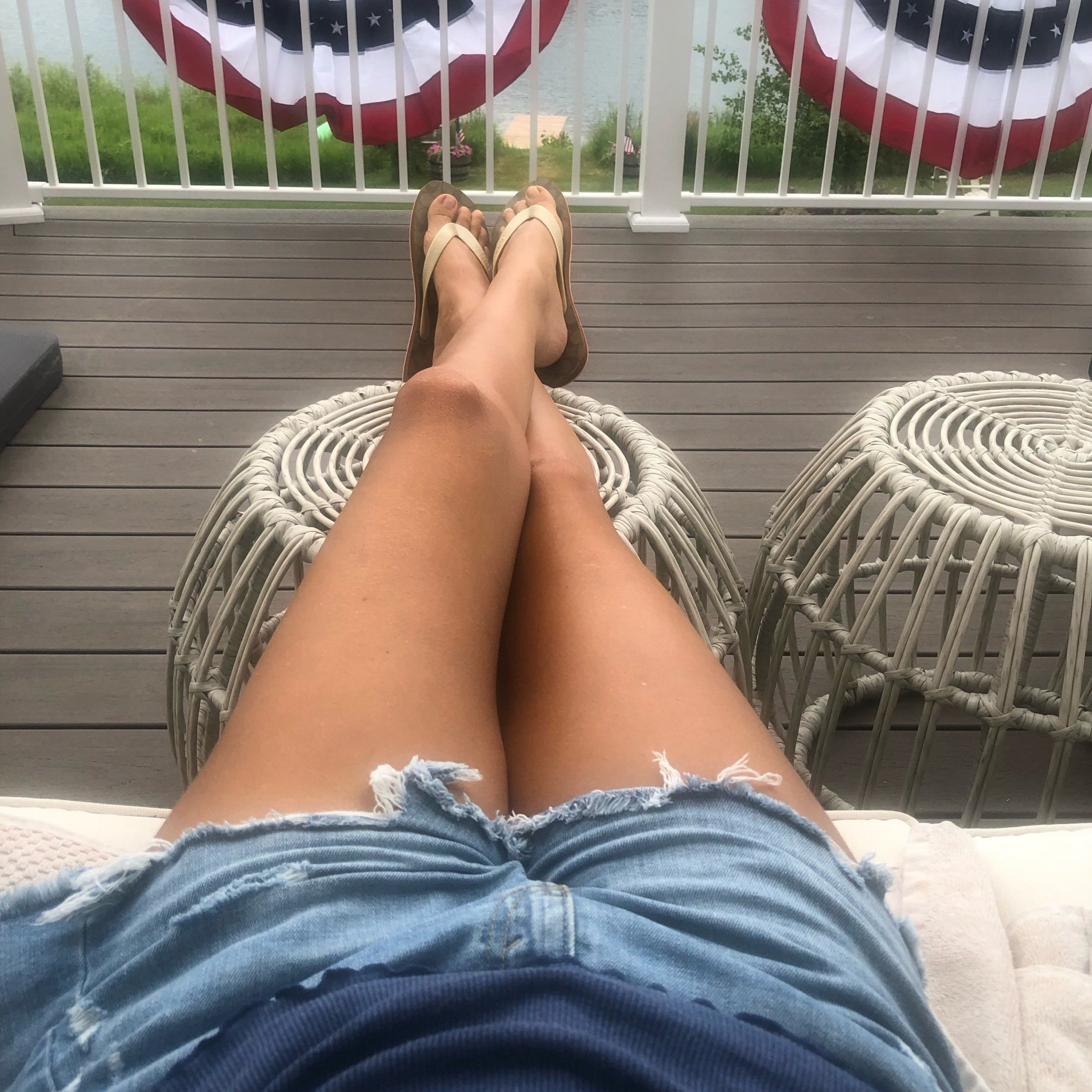 Amy outside on deck legs up on stool cut off denim shorts. Lake in background with american flags