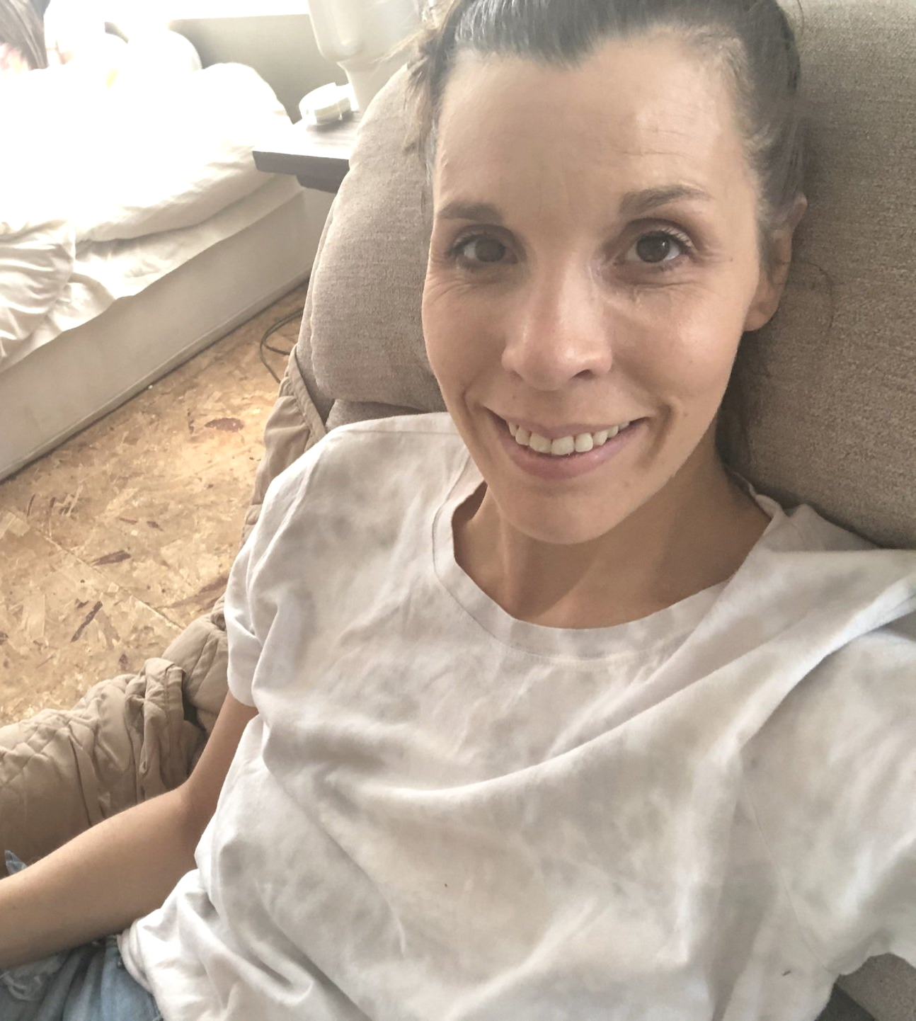 Amy smiling in recliner. Hair pulled back. Looking very thin and sick colored skin. No carpet flooring and bed on floor behind her