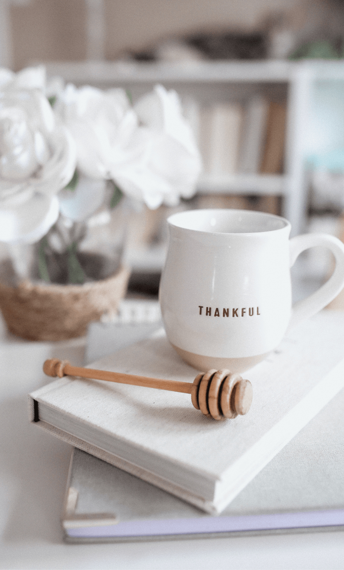 Notebook and journal stacked on top of each other with cup that has THANKFUL written on it. Also has a honey dipper next to it on the journal. All are sitting on a counter with a white flowered plant contrasted blurry in the background.