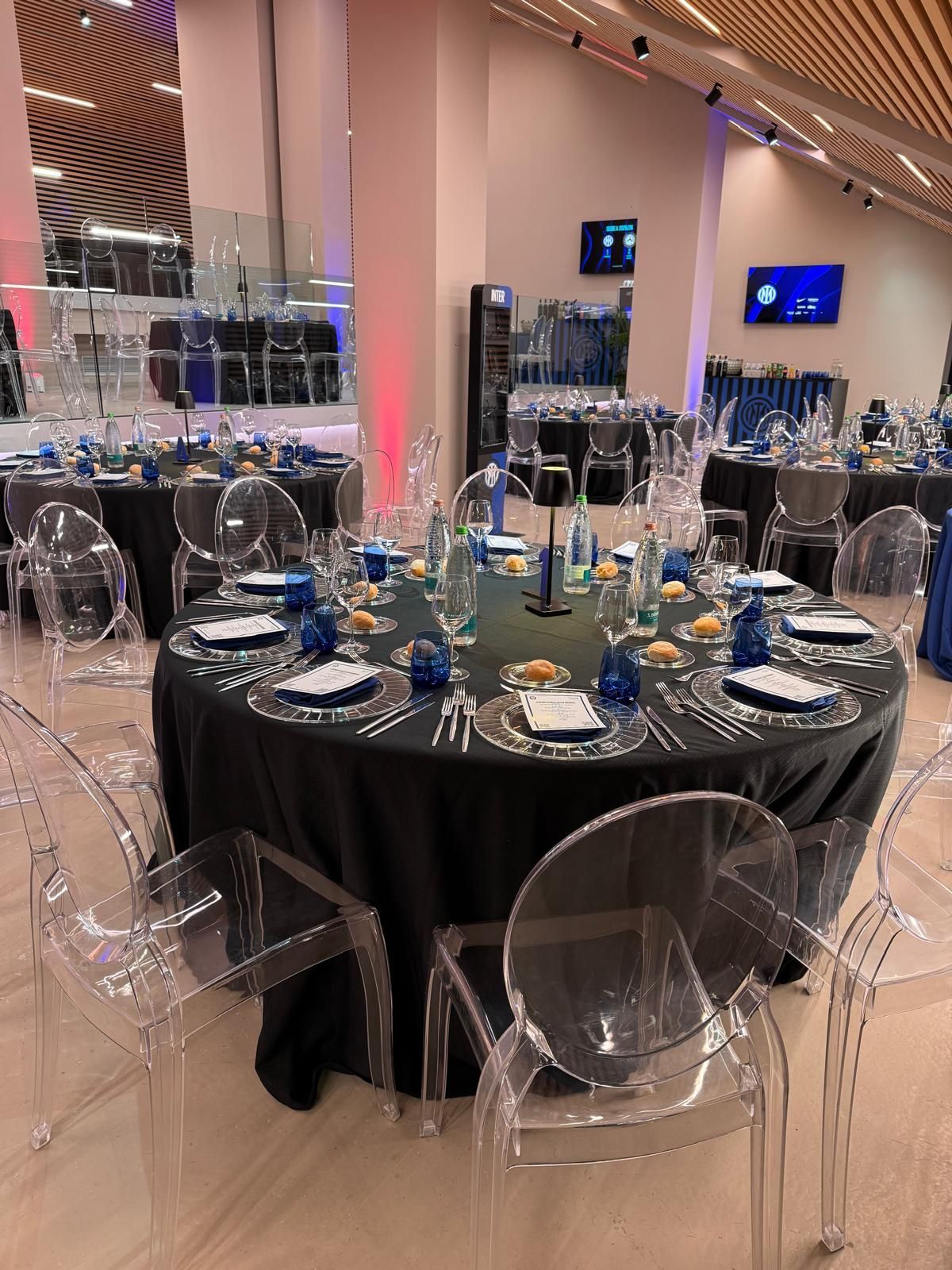 Tavoli rotondi apparecchiati per un evento formale, con tovaglie nere, sedie trasparenti e dettagli blu in un ambiente moderno.