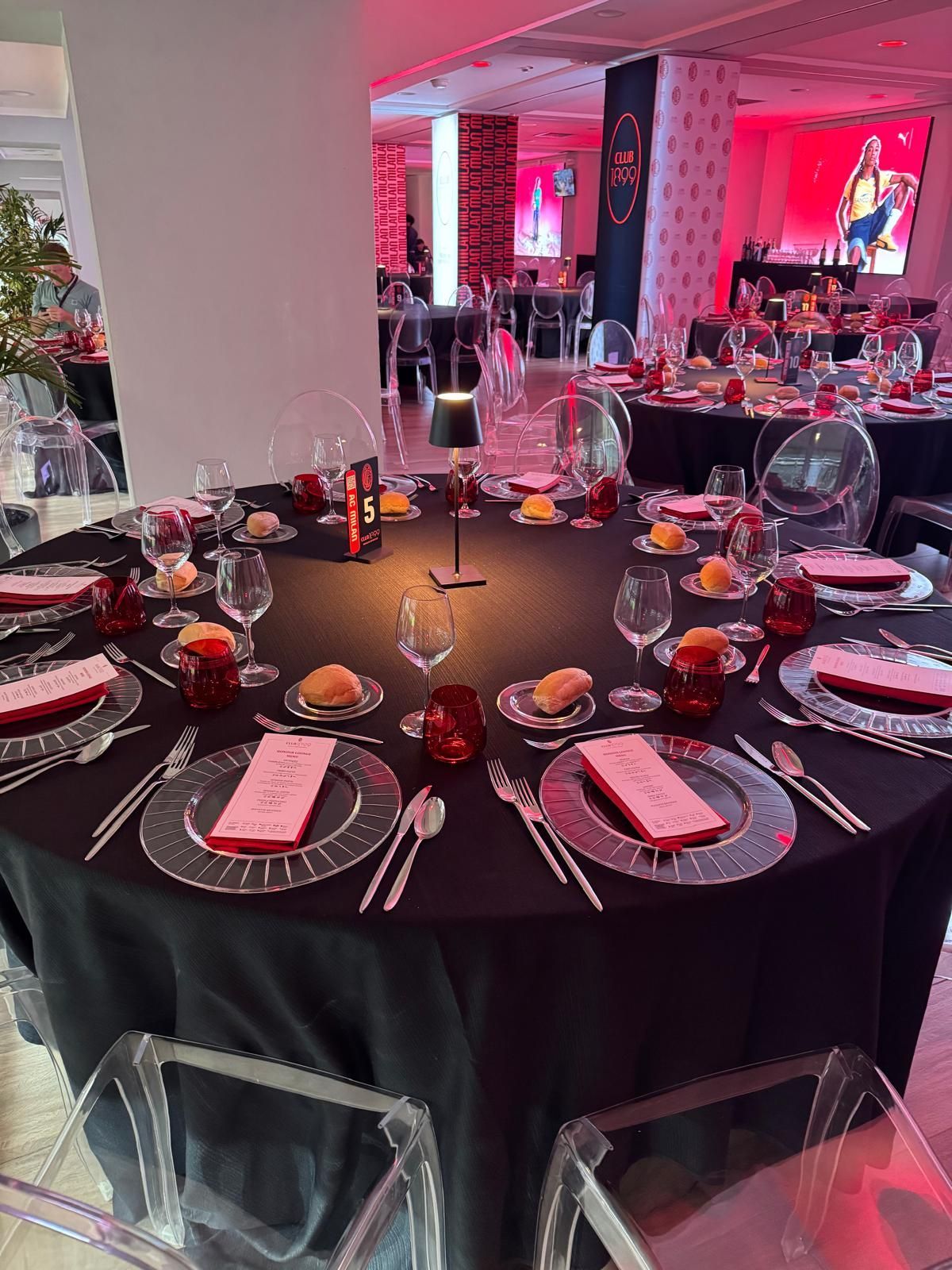 Elegante tavolo rotondo apparecchiato per una cena formale. Tovaglia nera con dettagli rossi e argento, sedie trasparenti e illuminazione rossa.