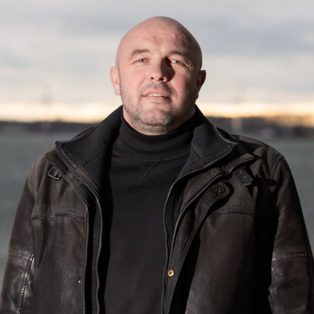 Bald man in a black turtleneck and leather jacket, outdoors with a neutral background.