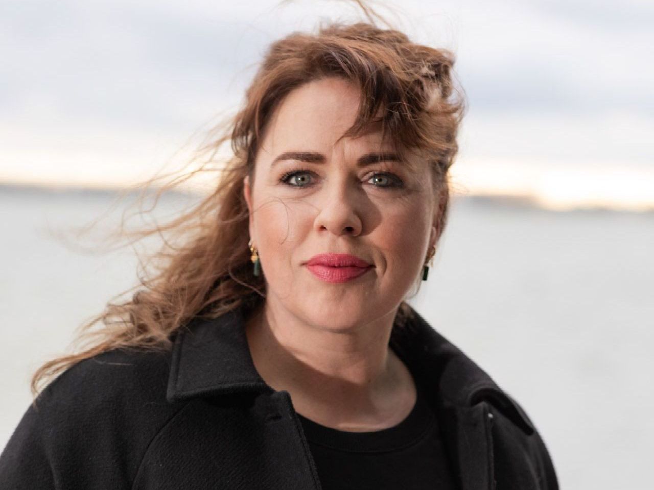 Woman with auburn hair and red lipstick, wearing a black coat, looking at the camera, with a blurred ocean background.