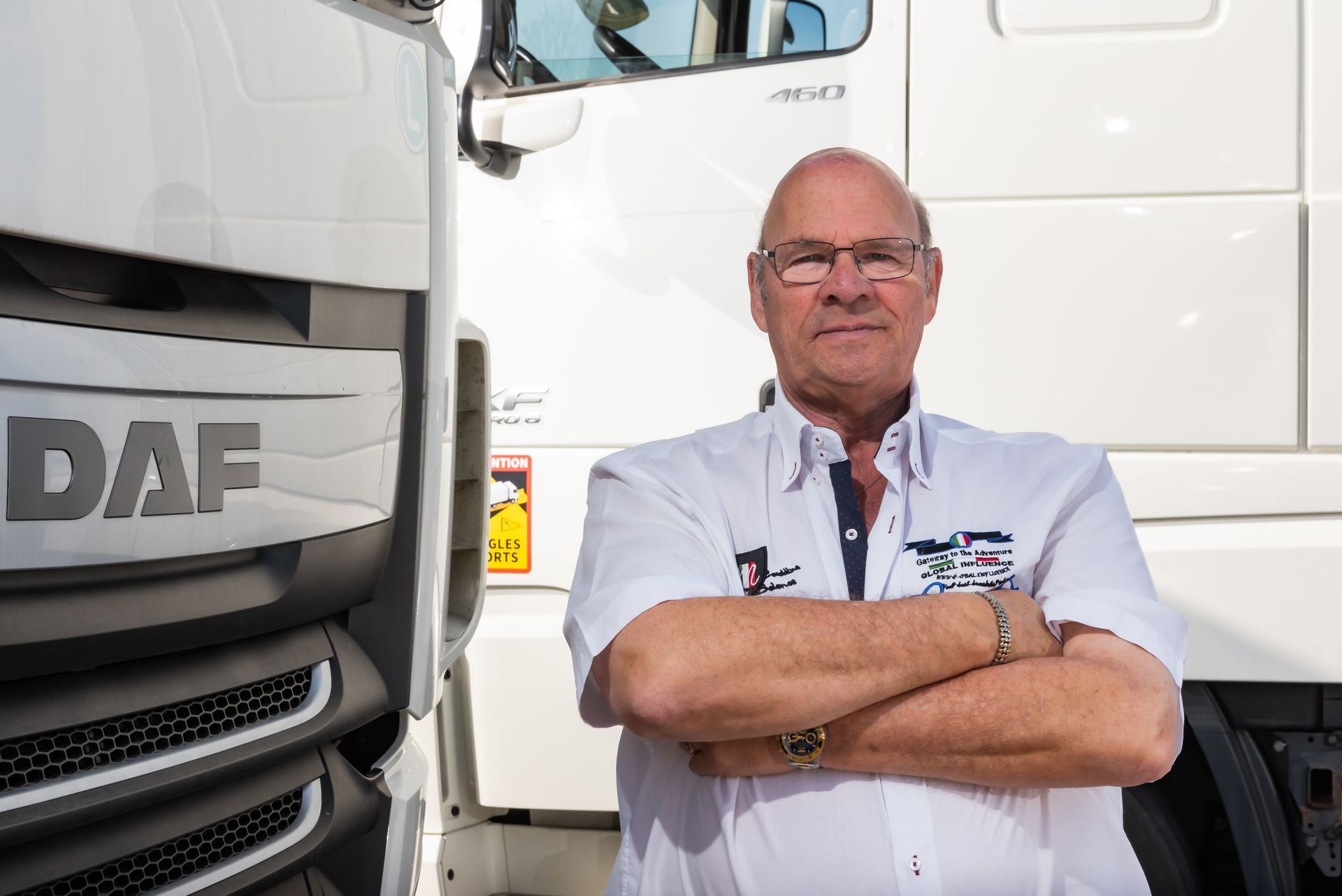 Man in white shirt with arms crossed standing next to a white DAF truck.