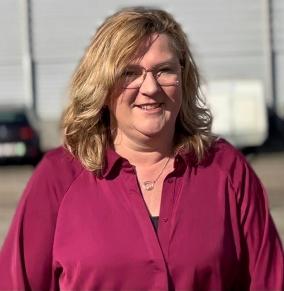 Woman with blonde hair and glasses, wearing a burgundy shirt, smiles outdoors.