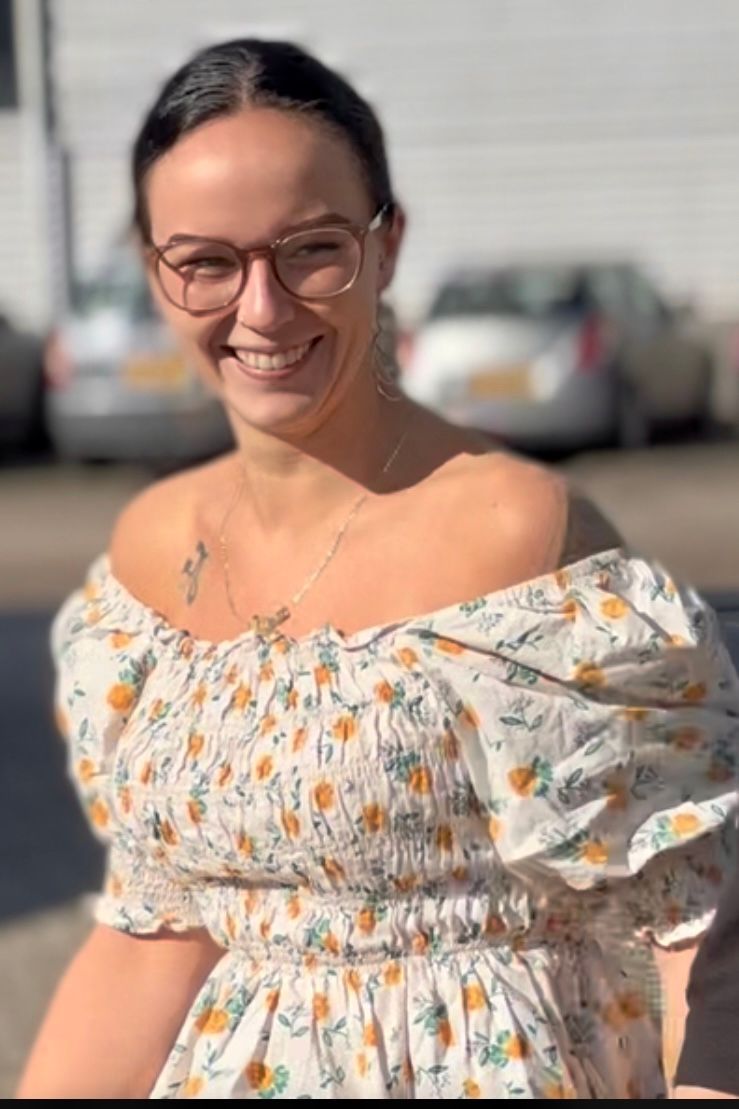 Woman with glasses smiles, wearing a white off-the-shoulder dress with yellow floral print, outdoors with cars blurred in the background.
