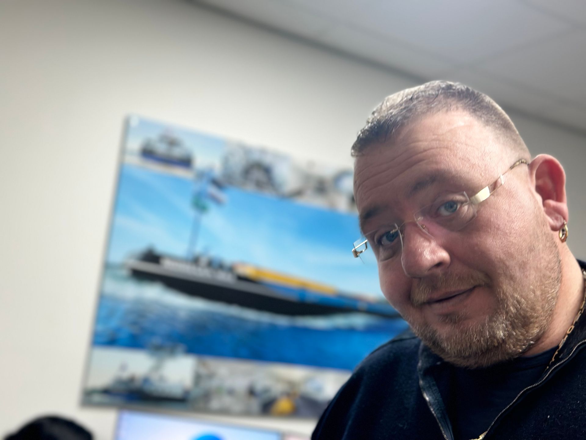 Man with glasses and a beard in front of a boat image. He's smiling, possibly in an office setting.