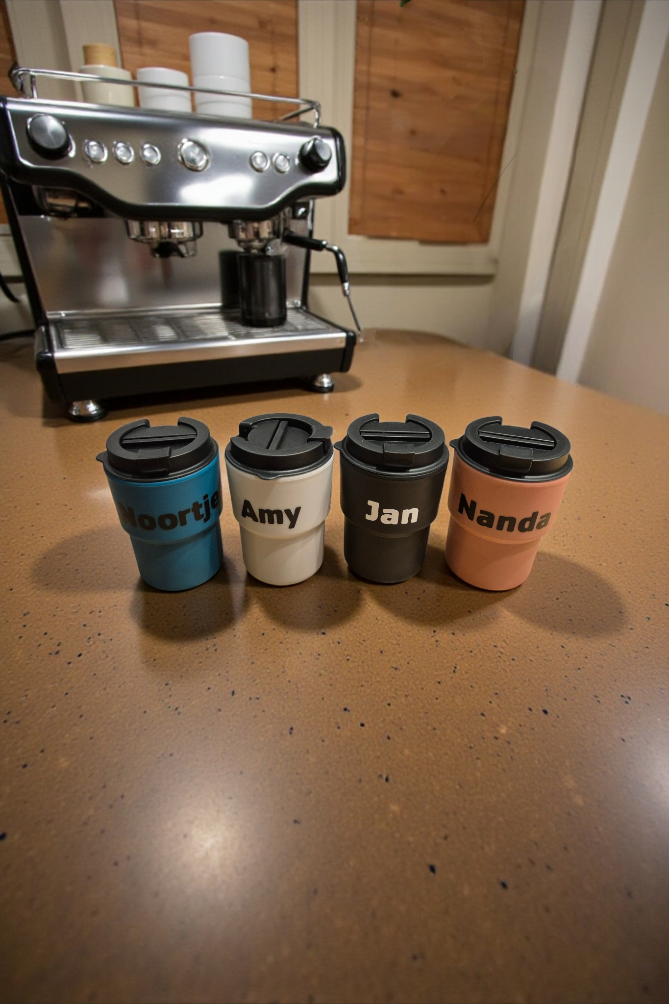 Op een tafel, voor een espressomachine, staan vier koffiemokken met deksels en een etiket erop.