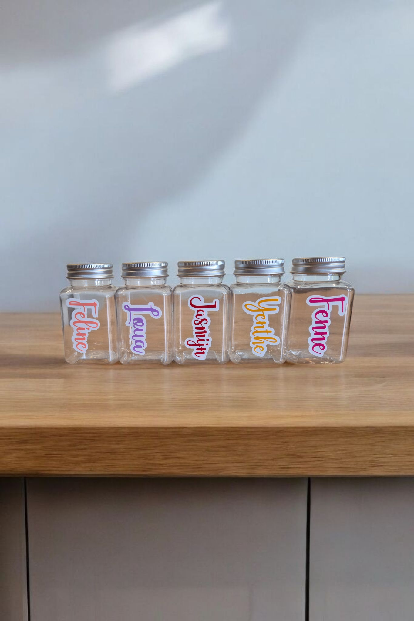 Vijf kleine plastic potjes met zilveren deksels, elk voorzien van een ander naamlabel in kleurrijk schrift.