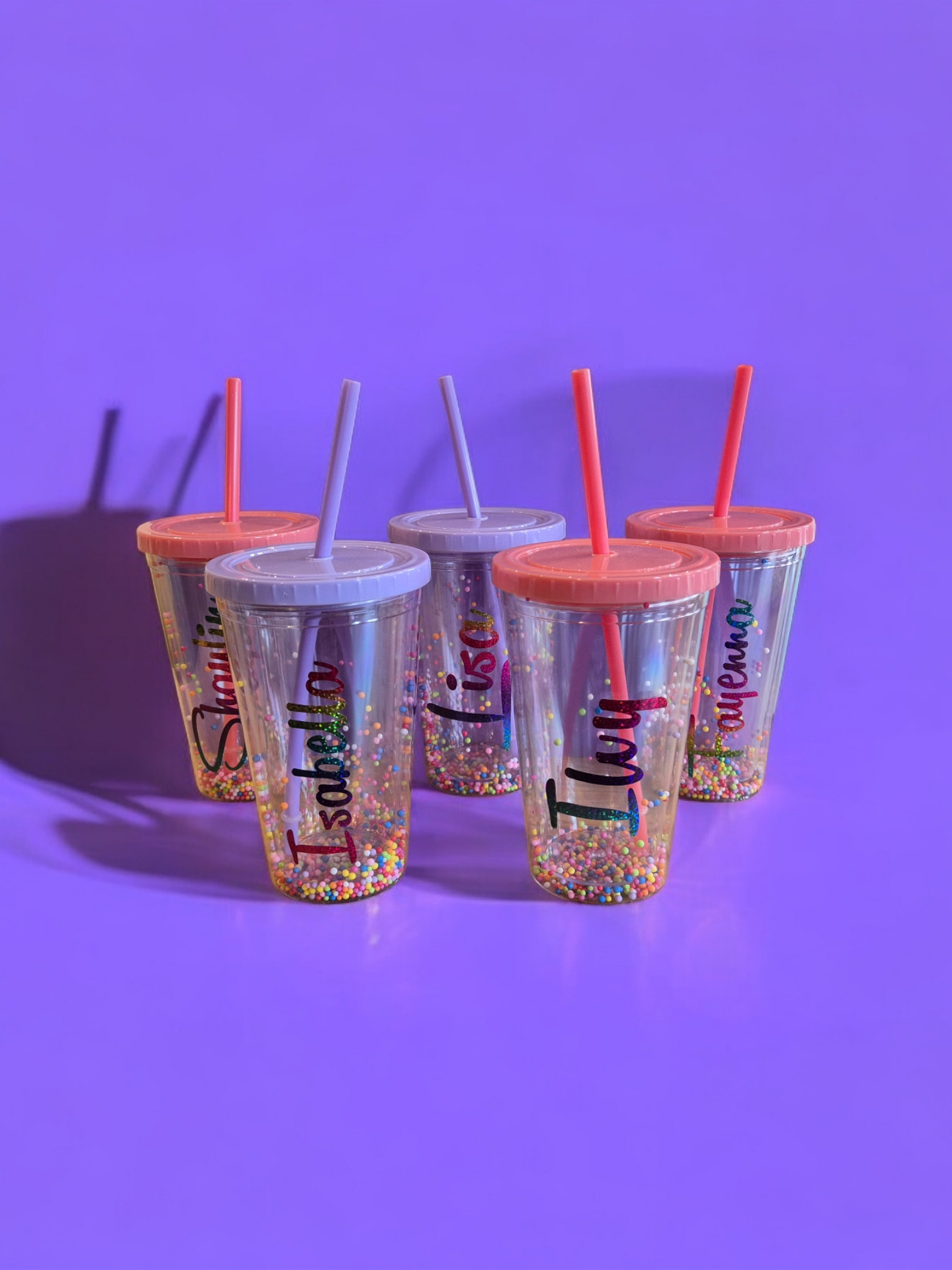Vijf gepersonaliseerde, met glitter gevulde drinkbekers met rietjes op een paarse ondergrond. Namen zijn erop gedrukt.