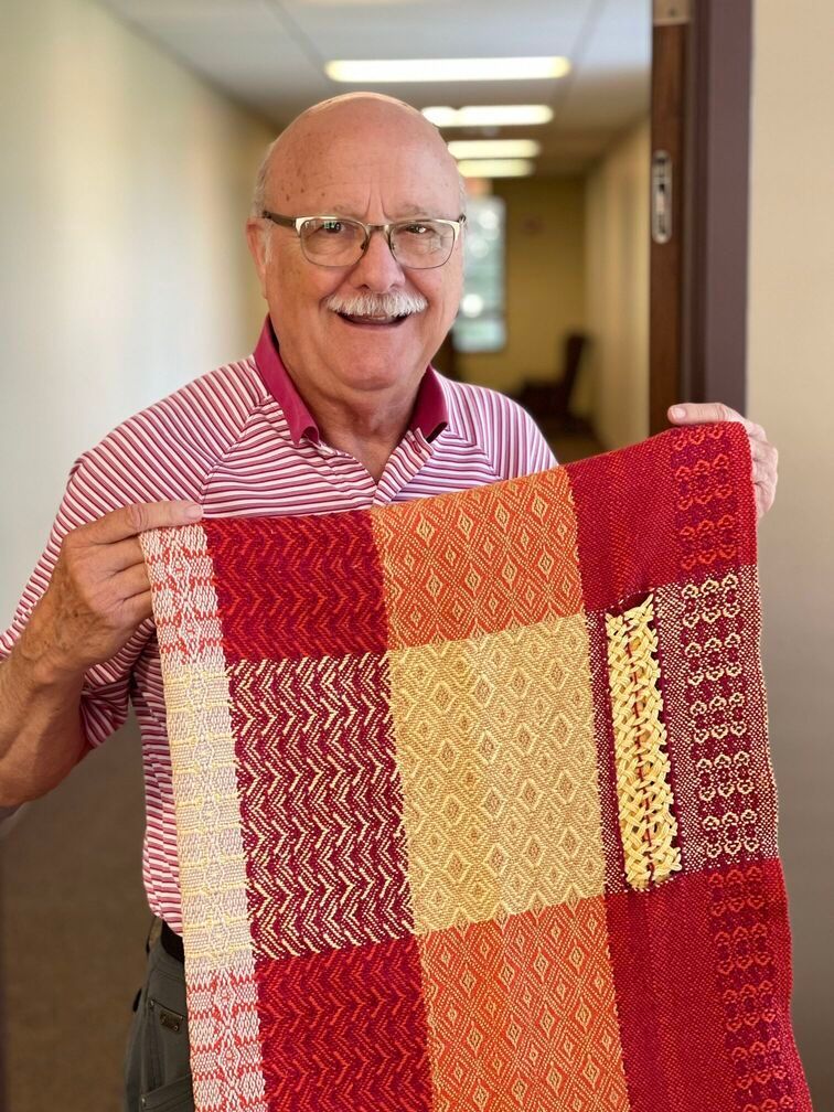 A male weaver's guild member  holding a red and yellow woven blanket in a hallway.
