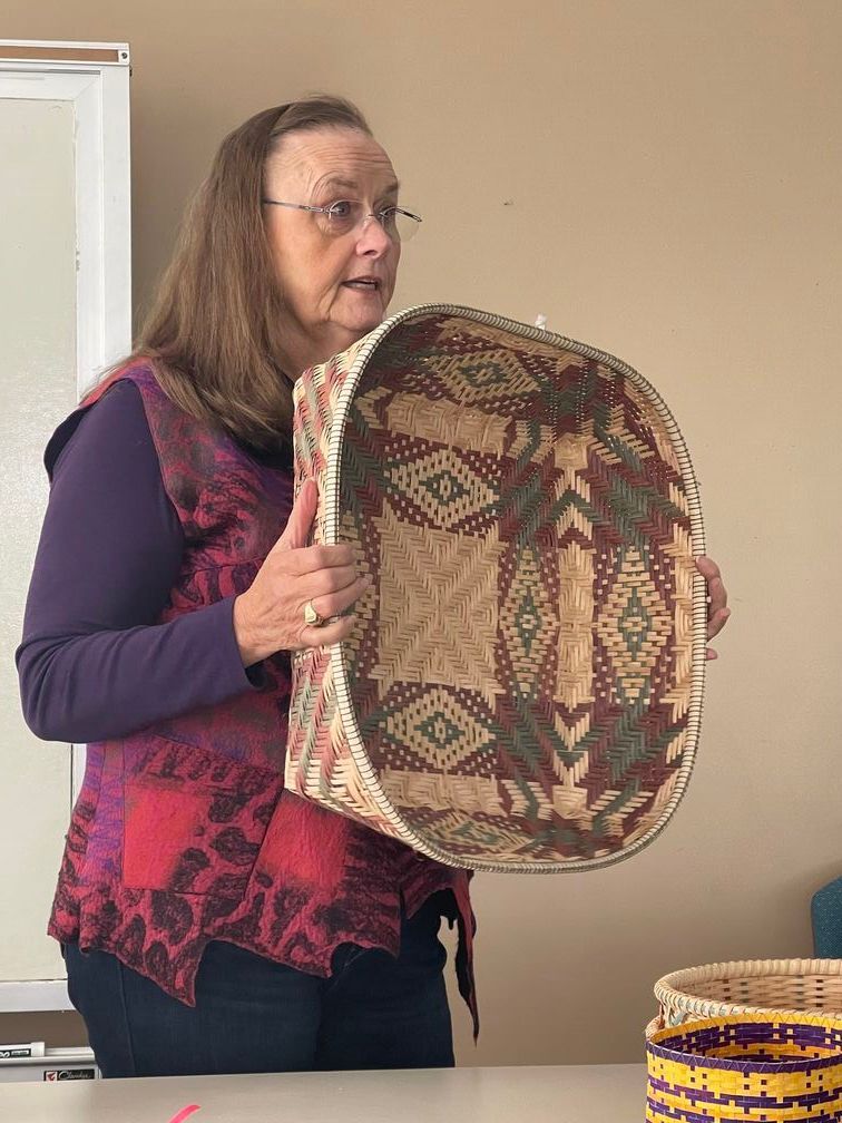 A weaver's group member in a purple vest holding a hand woven basket in her hands.