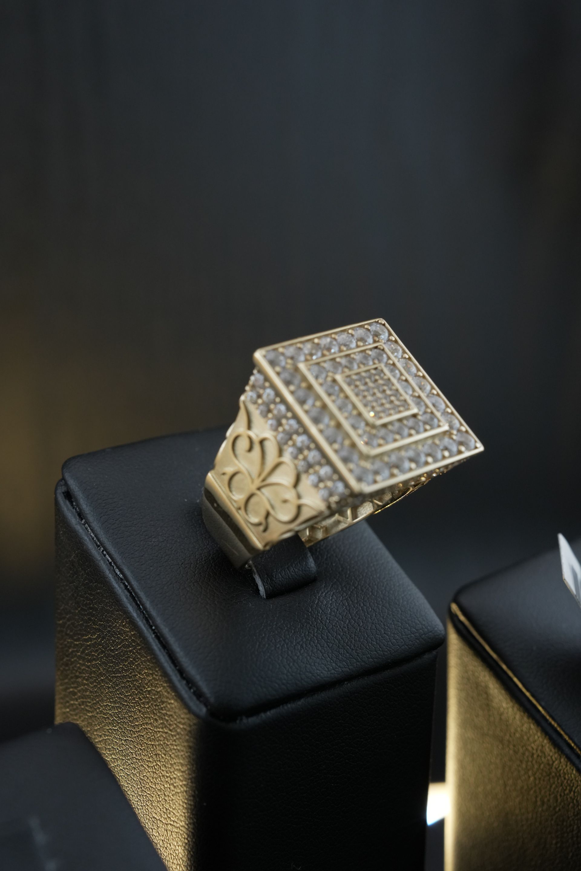 Gold ring with square diamond-encrusted top displayed on a black stand against a dark background.