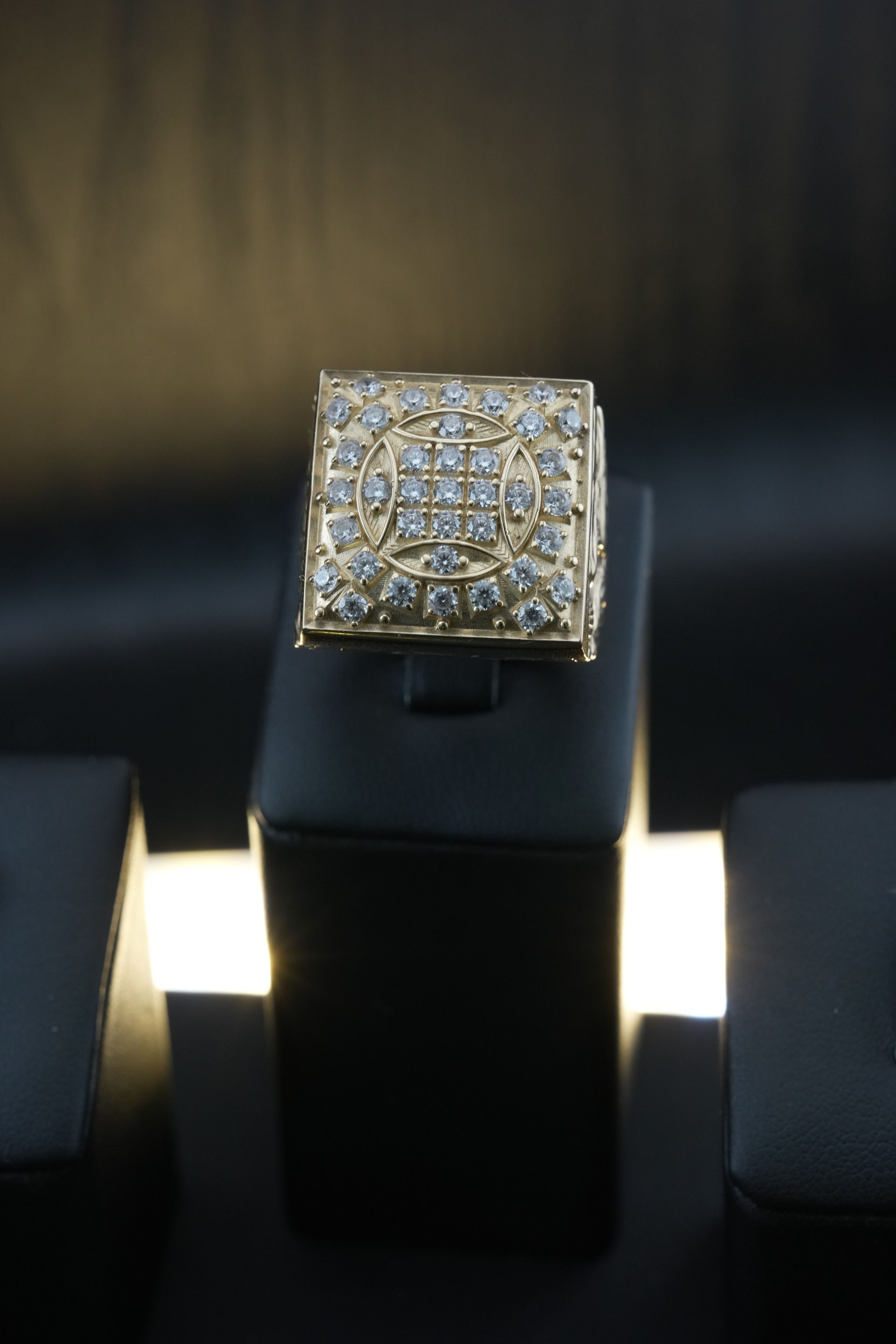 Square gold ring with many small diamonds on black display stand.